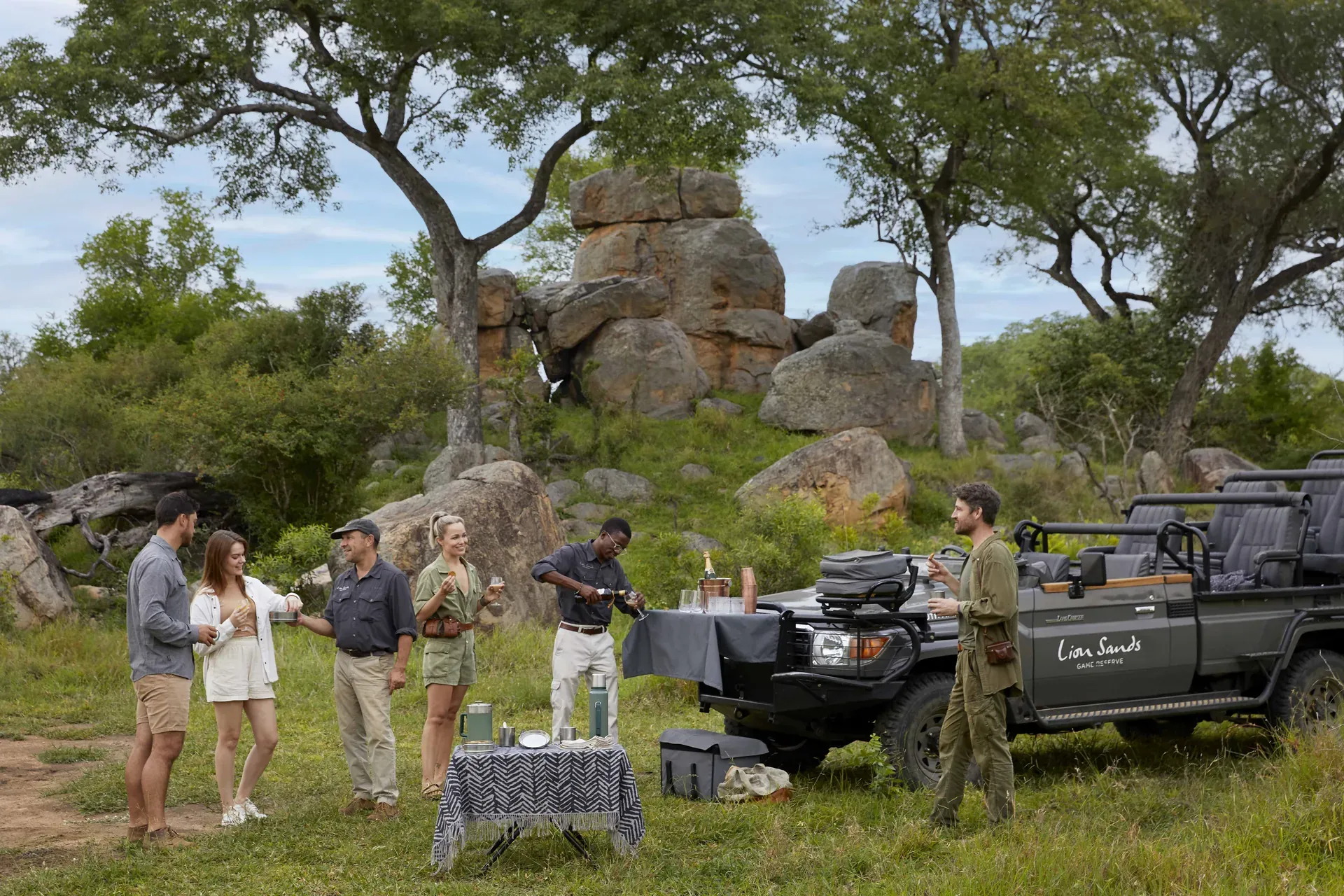 Outdoor safari setting with a dining table and lounge chairs under a shady tree in a grassy bush landscape